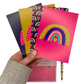 A hand holding a set of 4 rainbow postcards. The postcards are very bright and bold, in magenta, navy blue and mustard.