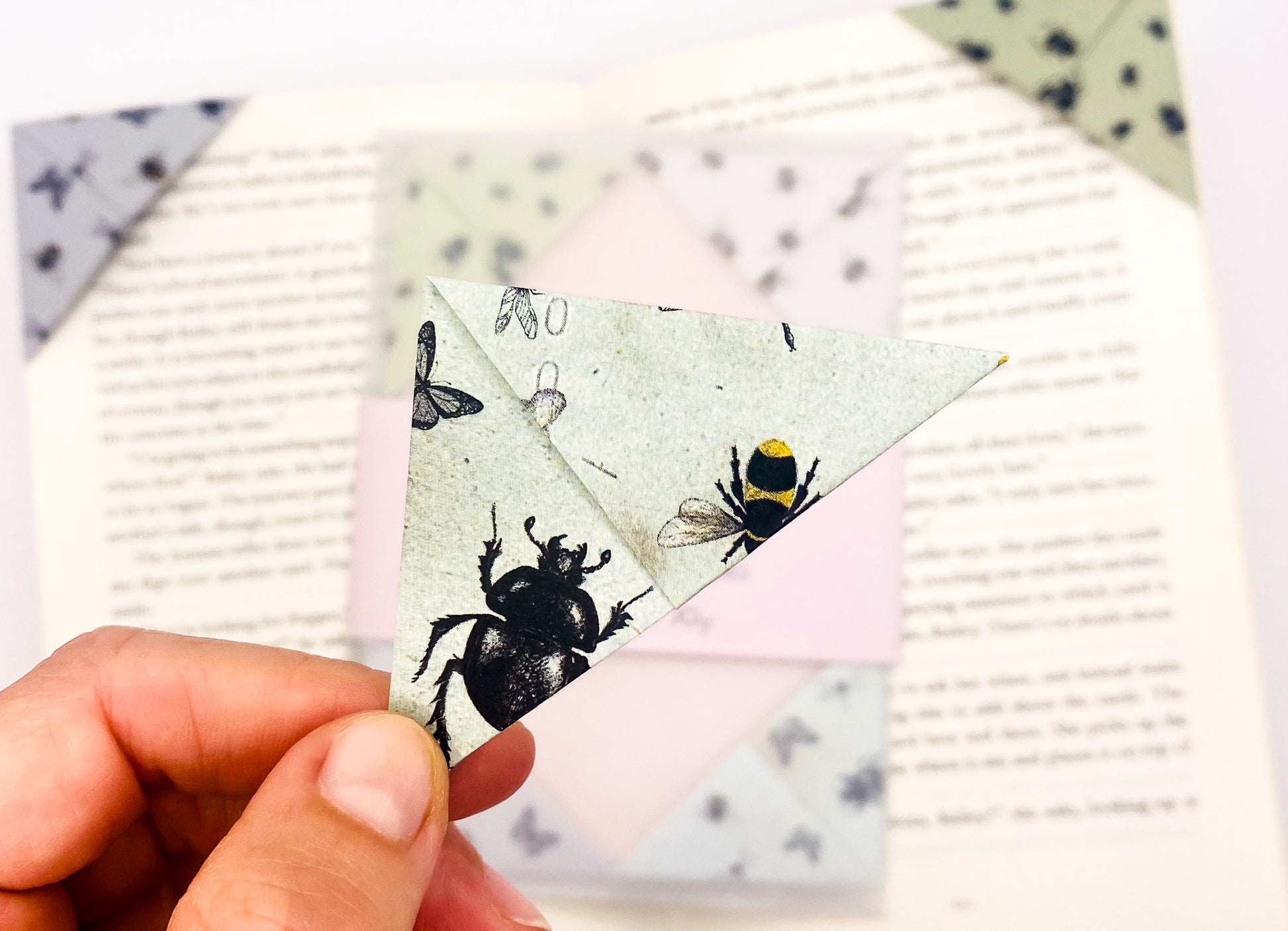 A close up of a hand holding an origami insect bookmark with a book in the background