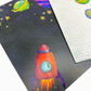 Close up image of two postcards in the space collection. The postcard in the foreground features a navy background with stars, planets and a red rocket.