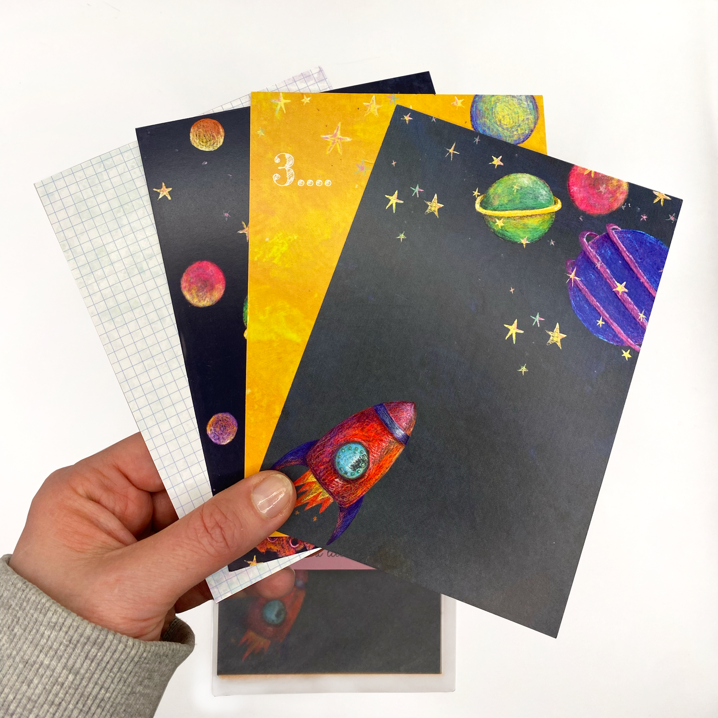 A hand holding 4 space themed postcards. The main postcard has a navy background with stars, planets and a red rocker